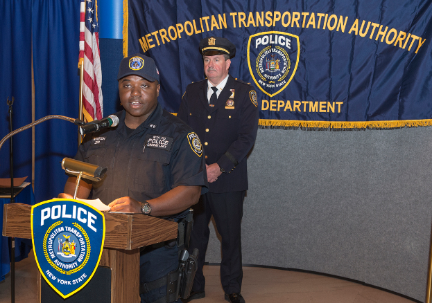 Zwei Polizisten vor einem Rednerpult stehend, einer hält Papiere und spricht in ein Mikrofon, mit einer Fahne, einem Tisch und einer Tafel mit Symbolen und Text im Hintergrund.