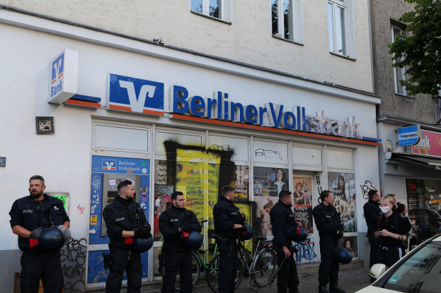 Eine Gruppe von Polizisten steht vor einer Berliner Volksbank, einige halten Helme, mit einem Auto auf der rechten Seite, einem Baum auf der linken Seite und Gebäuden mit Fenstern und Texttafeln im Hintergrund.