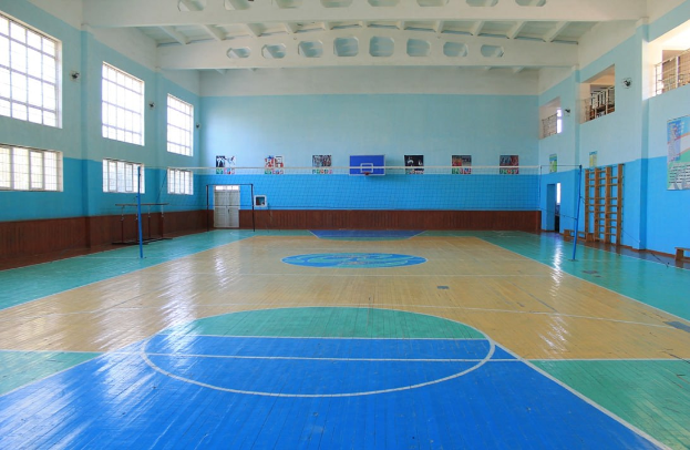 Große Turnhalle mit blau-weißem Boden, einem Basketballfeld, einem Netz mit Pfosten, einer Tür, Fenstern, Plakaten an den Wänden, einem Geländer und einem Dach.