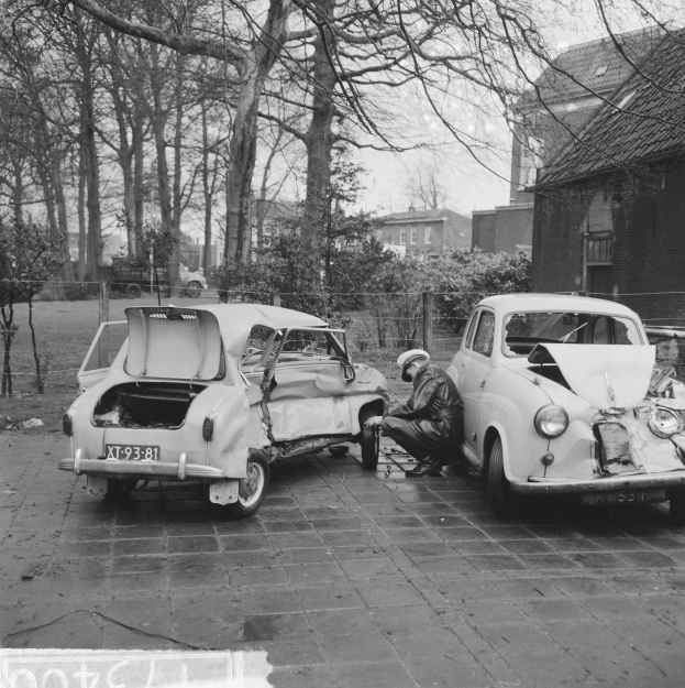 Zwei Autos bei einem Unfall an der Straßenseite beteiligt, mit einer Person im Vordergrund sitzend und Bäumen, Gebäuden, Pflanzen und einem Zaun im Hintergrund, dargestellt in Schwarz-Weiß.