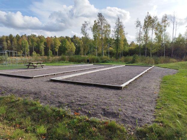 Eine grüne Wiese mit einem Picknicktisch in der Mitte, umgeben von Bäumen und einem klaren blauen Himmel, mit einem Spielplatz mit Pfählen und einer Bank links.