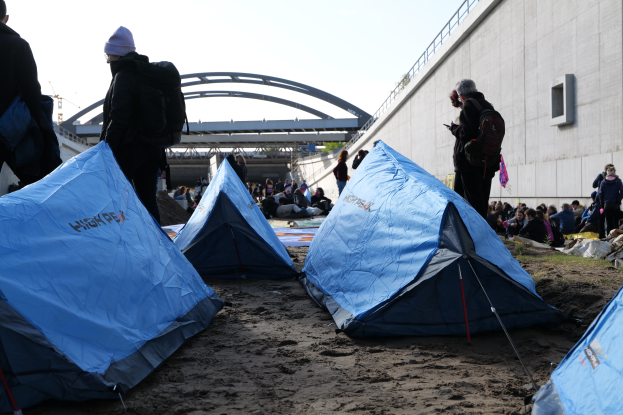 Eine Gruppe von Menschen sitzt auf einem sandigen Strand in der Nähe von Zelten, mit einer Wand auf der rechten Seite und einer Brücke im Hintergrund, die besorgt aussieht.