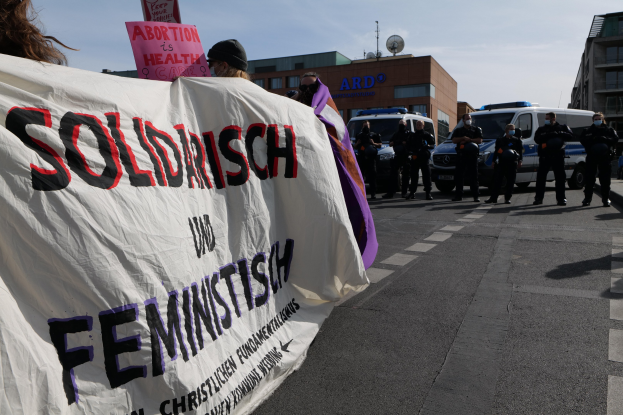 Eine Gruppe von Menschen, die eine Straße entlanggehen und ein Transparent mit der Aufschrift 'Solidarität und Feminismus' tragen, mit parkenden Fahrzeugen, Gebäuden, einer Satellitenschüssel und einem klaren blauen Himmel im Hintergrund.