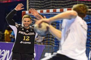 Zwei Männer spielen Handball vor einem Tor mit Netz und einer Fahne mit Text im Hintergrund und einigen Zuschauern in einer Arena.