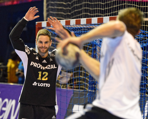 Zwei Männer spielen Handball vor einem Tor mit Netz und einer Fahne mit Text im Hintergrund und einigen Zuschauern in einer Arena.