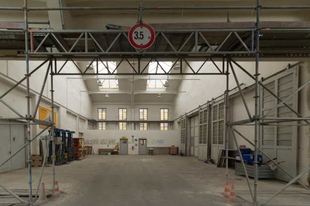 Großer Lagerraum mit ausgedehnten Gerüsten, Verkehrskegeln, Metallstangen, einem Schild, Maschinen, einer Tür, Fenstern, Wänden, Deckenlampen und einem Dach.