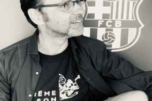 Ein Mann mit Brille und schwarzem Hemd lächelt vor einem schwarz-weißen FC Barcelona-Logo.