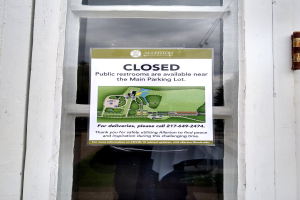 Glasstür mit einem Schild "Geschlossene öffentliche Toiletten verfügbar in der Nähe des Hauptparkplatzes" und einem unklaren Objekt auf der rechten Seite.