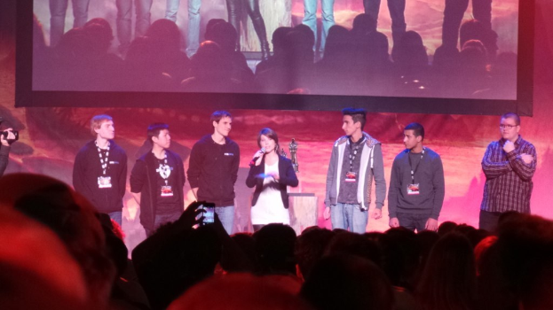 Eine Gruppe von Menschen auf einer Bühne, einer hält ein Mikrofon, mit einigen Zuschauern davor und einem Bildschirm im Hintergrund.