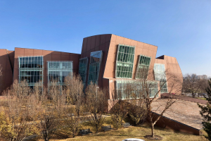 Großes mehrstöckiges Gebäude mit zahlreichen Fenstern, umgeben von Bäumen, Bänken, Gras und einem Laternenmast, vor einem klaren blauen Himmel mit anderen Gebäuden im Hintergrund.