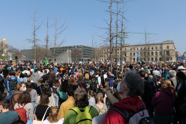 Eine große Menge Menschen steht vor einem Gebäude mit Fenstern, Bäumen und einem klaren Himmel, viele halten Schilder und tragen Taschen, was auf eine Klimawandel-Demonstration hinweist.