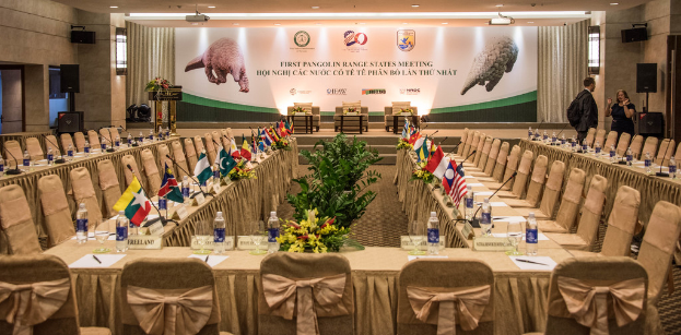 Eine Bühne mit Stühlen, Mikrofonen, Flaggen, Flaschen auf Tischen und einigen stehenden Menschen sowie einem Plakat.