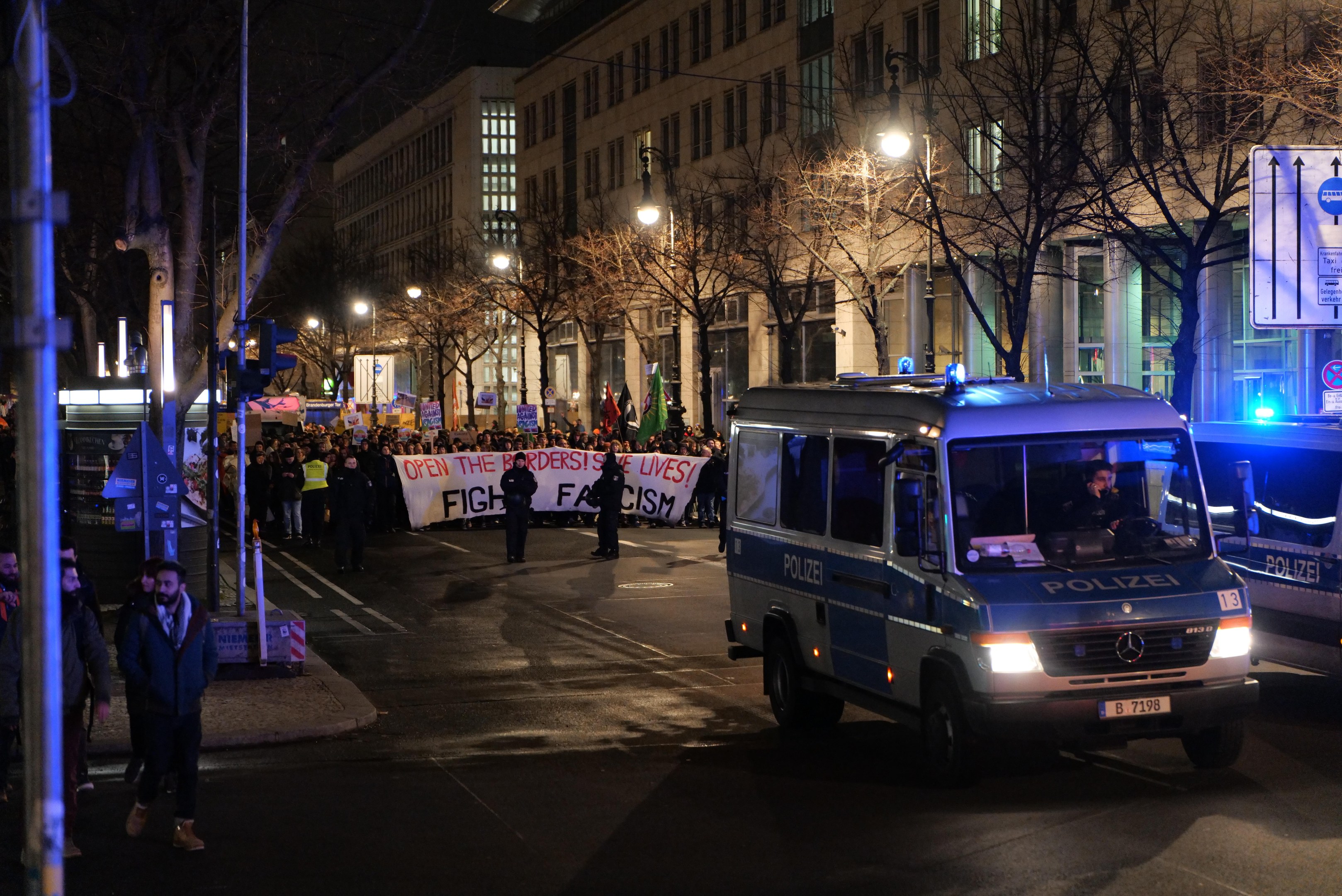 Eine Gruppe von Menschen, die nachts eine Straße entlanggehen, mit einem geparkten Polizeiwagen im Vordergrund, umgeben von Bäumen und Gebäuden und einem Banner im Hintergrund.