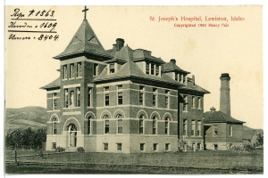 Ein Schwarz-Weiß-Foto des St. Joseph-Krankenhauses in Lewiston, Idaho, das ein großes Gebäude mit Fenstern, Treppen mit Geländern, einen Schornstein auf der rechten Seite und Text oben zeigt, mit Bäumen, Hügeln und dem Himmel im Hintergrund.