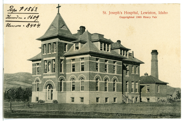 Ein Schwarz-Weiß-Foto des St. Joseph-Krankenhauses in Lewiston, Idaho, das ein großes Gebäude mit Fenstern, Treppen mit Geländern, einen Schornstein auf der rechten Seite und Text oben zeigt, mit Bäumen, Hügeln und dem Himmel im Hintergrund.