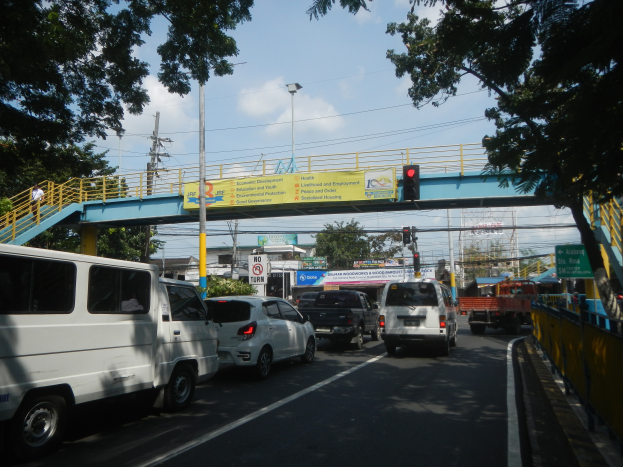 Eine belebte Straße mit Autos und Lastwagen, eine Brücke mit Geländern und Treppen, Laternen, Ampeln, Schilder mit Text, Bäume, Gebäude und ein bewölkter Himmel.