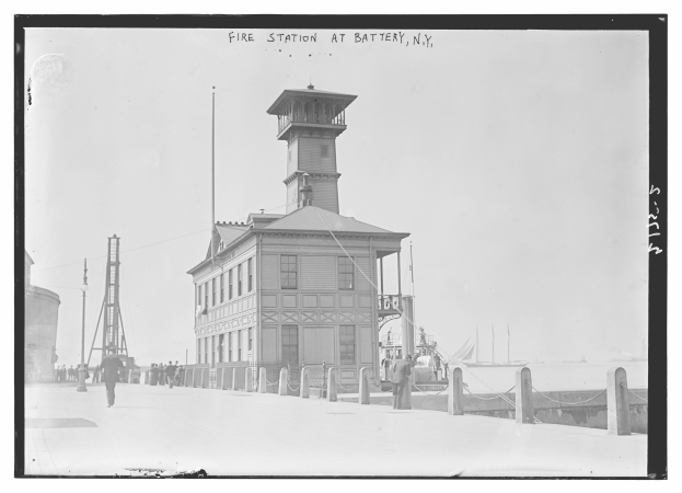 Ein Schwarz-Weiß-Foto einer Feuerwache an der Battery, New York, mit einem Gebäude, Menschen, Mästen, einem Zaun, Booten auf dem Wasser und einem bewölktem Himmel.