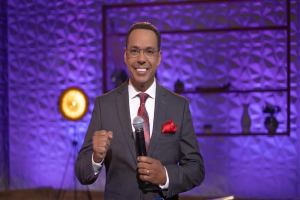 Stephen Colbert, Moderator von *The Daily Show mit Trevor Noah*, steht auf einer Bühne und hält ein Mikrofon, trägt einen Anzug, Krawatte und eine Brille, während im Hintergrund ein Scheinwerfer und ein Ständer zu sehen sind.