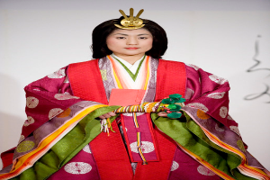 Eine Frau in einem traditionellen japanischen Kimono, das ein leuchtendes rotes und grünes Kleid mit kunstvollen Mustern zeigt, steht selbstbewusst auf einem Laufsteg, ihr Haar ordentlich zu einem Knoten gebunden.