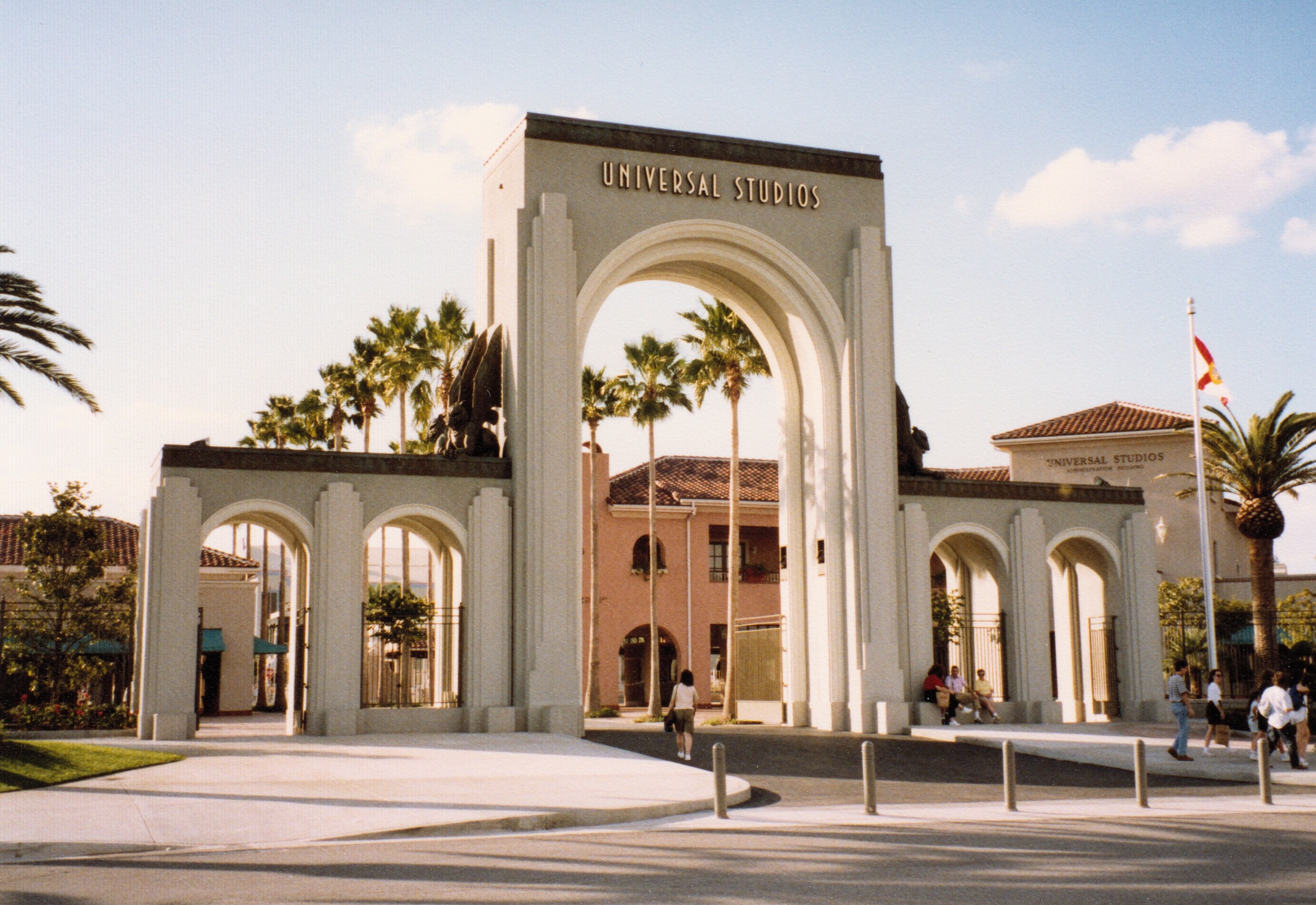 Eingang zu Universal Studios Hollywood mit einem Tor, einer Statue, Menschen, Bäumen, Gebäuden, einer Fahnenstange und einem bewölkten Himmel.