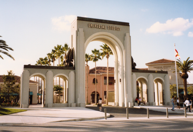 Eingang zu Universal Studios Hollywood mit einem Tor, einer Statue, Menschen, Bäumen, Gebäuden, einer Fahnenstange und einem bewölkten Himmel.