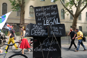 Eine Gruppe von Menschen geht auf der Straße mit Fahrrädern, einige tragen Taschen und einer hält eine Fahne, im Hintergrund gibt es Bäume, ein Gebäude mit Fenstern und ein Schild mit der Aufschrift "Stoppt Abschiebung".