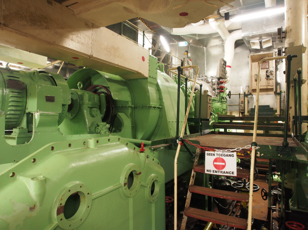 Ein großer grüner Generator in einem Gebäude umgeben von verschiedenen Maschinen, einer Leiter, einem Schild, Rohren, Lampen und anderen Gegenständen.