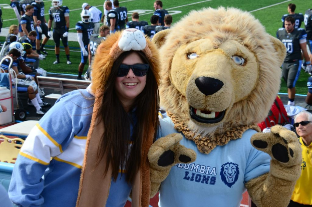 Eine Frau steht in der Nähe einer Person in einem Löwenkostüm, mit Spielern auf dem Boden dahinter, einigen Menschen auf einer Bank links und Stühlen neben der Bank.