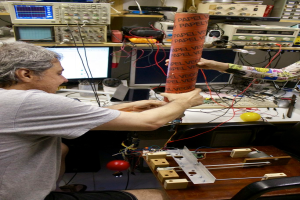 Ein Arbeitsplatz mit Maschinen, Kabeln, einem Computerbildschirm, einem Kugelschreiberhalter, Menschen und einem Stressball.