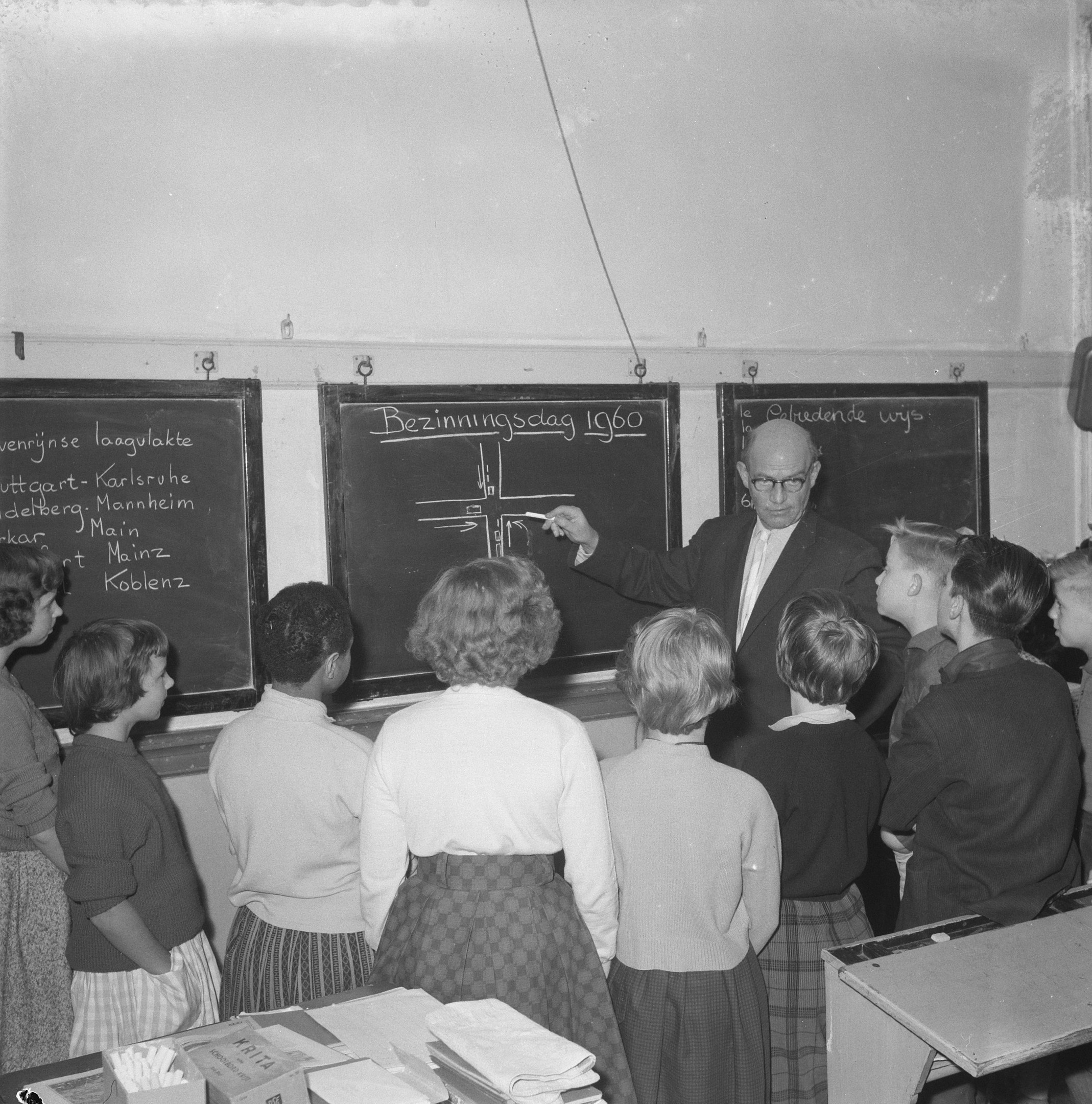 Schwarzes und weißes Foto eines Lehrers, der vor einer Tafel steht und einen Marker hält, während Kinder an einem Tisch mit Büchern und Gegenständen sitzen; ein an der Wand befestigter Lautsprecher ist auf der rechten Seite zu sehen.