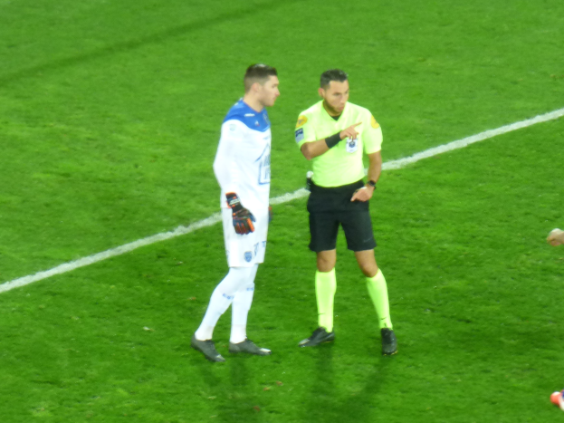 Ein Schiedsrichter in einem gelben Hemd und schwarzen Shorts spricht mit einem Fußballspieler in einer weißen und blauen Uniform auf dem Feld, beide tragen Turnschuhe.