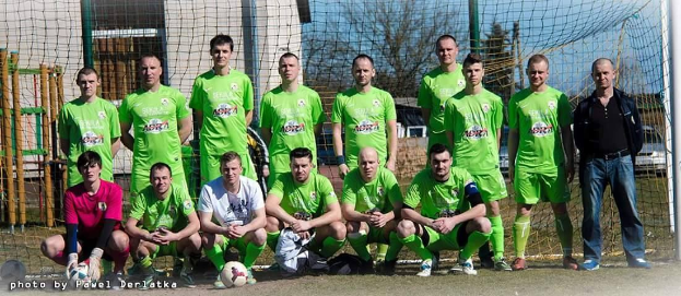 Gruppe von Männern in grünen T-Shirts, die auf einem Füßballfeld mit einem Ball und einem Tor stehen, mit Bäumen und Gebäuden im Hintergrund.