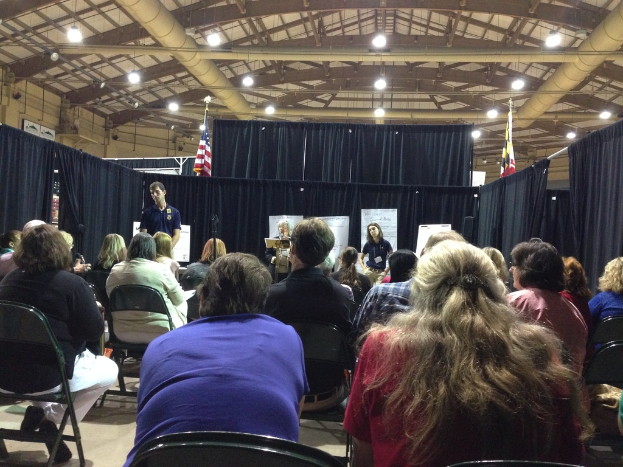 Eine Gruppe von Menschen, die auf Stühlen sitzen, mit einer Person, die an einem Rednerpult steht, vor einem Hintergrund aus Banner, blauen Vorhängen, Flaggen und Deckenleuchten.