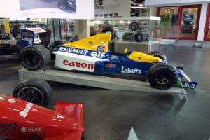 Eine Museumausstellung mit verschiedenen Rennwagen, darunter ein Renault F1-Auto, mit Besuchern, die die Ausstellungsstücke betrachten, einem Textschild an der Wand und einer beleuchteten Decke sowie Glastüren auf der rechten Seite.