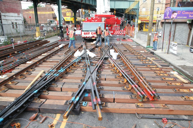 Arbeiter in Jacken und Mützen bauen Eisenbahnschienen, mit einem Fahrzeug, das mit Materialien beladen ist, und einem Schuppen mit Pfählen im Hintergrund und einem Geschäft auf der anderen Seite.