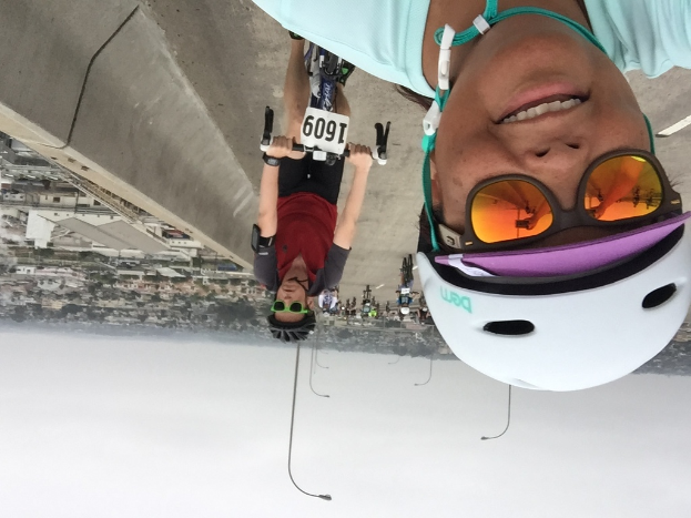 Eine Gruppe von Menschen, die auf Fahrrädern auf einem Weg fahren, mit Bäumen und Gebäuden auf der linken Seite, Laternenmasten dahinter und einem Himmel im Hintergrund.