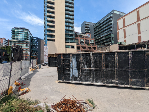 Ein großer Metallcontainer steht auf einem Parkplatz umgeben von Gebäuden, einem Metallzaun, Pflanzen, Bäumen, Fahrzeugen und Pfählen unter einem bewölkten Himmel, mit Text, der ein Bauprojekt in Toronto anzeigt.