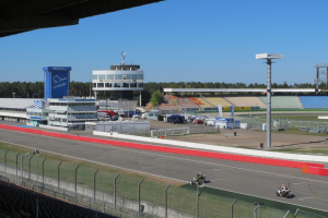 Eine Gruppe von Menschen auf Motorrädern auf einer Rennstrecke umgeben von einem Metallzaun, Gras und einem bewölkten Himmel.