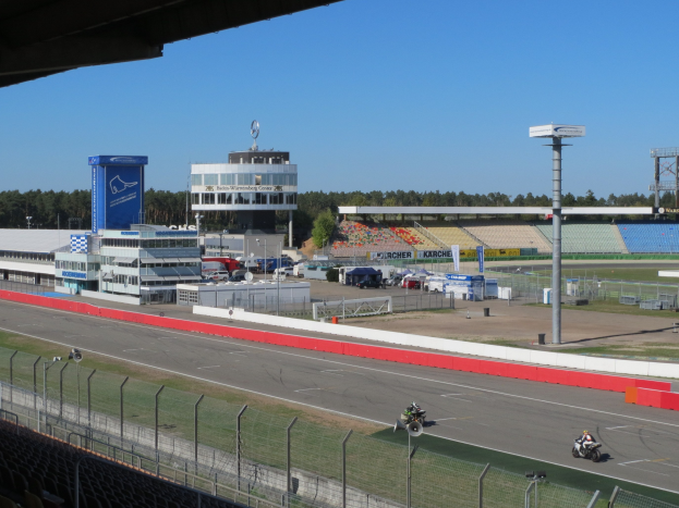 Eine Gruppe von Menschen auf Motorrädern auf einer Rennstrecke umgeben von einem Metallzaun, Gras und einem bewölkten Himmel.