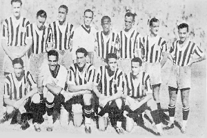 Ein Schwarz-Weiß-Foto einer Fußballmannschaft, wobei einige Spieler stehen und andere auf dem Boden sitzen, mit Text unten.