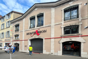 Eine Gruppe von Menschen mit Fahnen geht eine Straße vor einem Gebäude mit einer Schildertafel 'Montbron', einem Straßenschild und einer Banner mit Bildern und Text, unter einem bewölkten Himmel.