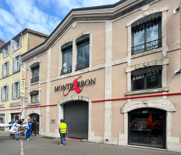 Eine Gruppe von Menschen mit Fahnen geht eine Straße vor einem Gebäude mit einer Schildertafel 'Montbron', einem Straßenschild und einer Banner mit Bildern und Text, unter einem bewölkten Himmel.