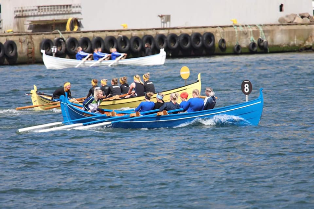 Eine Gruppe von Menschen in einem blauen und gelben Boot auf dem Wasser, die Paddel halten, mit einer Wand aus Reifen und einem Gebäude im Hintergrund, das eine Regatta vermuten lässt.