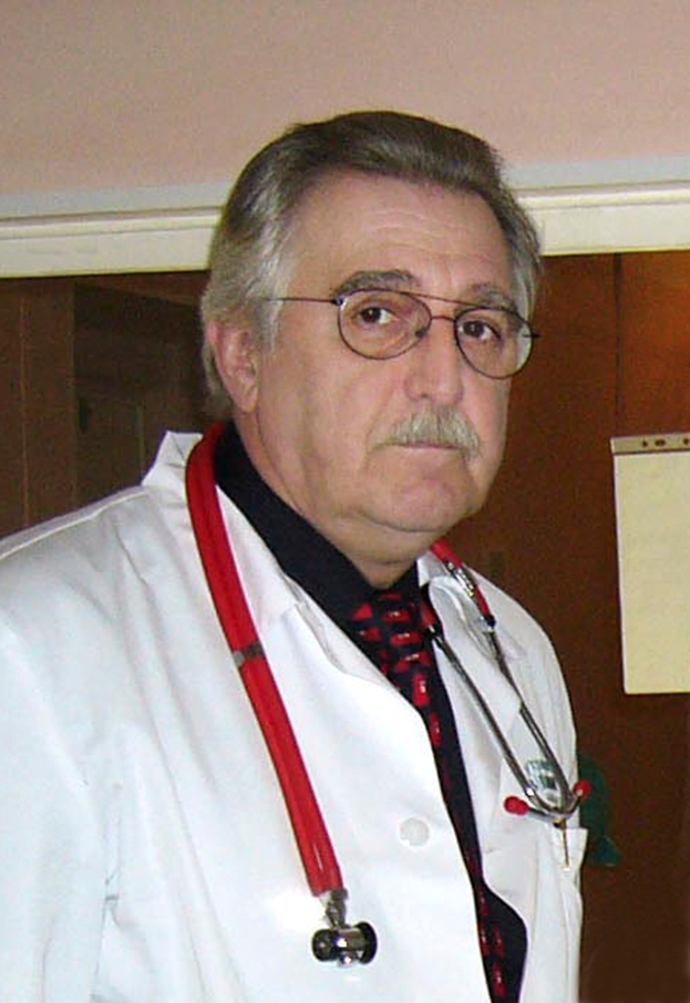 Ein Mann in einem weißen Kittel mit einem Stethoskop um den Hals steht vor einer Wand mit einem Blatt Papier daran und sieht ernst aus.