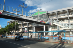Eine belebte Stadtstraße mit Fahrzeugen, einer Brücke, Strommasten, Laternen, Bäumen, Gebäuden mit Fenstern, Transparenten und einem bewölkten Himmel.