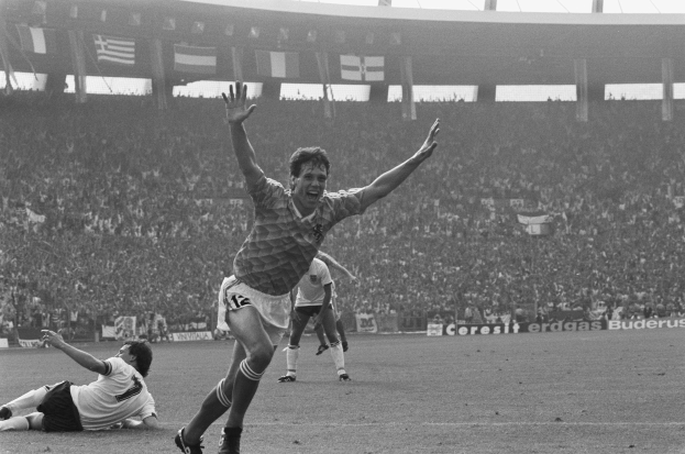 Ein Schwarz-Weiß-Foto eines Fussballspielers, der nach einem Tor jubelt, umgeben von Menschen auf dem Boden, mit Schildern, Fahnen und Pfählen im Hintergrund unter einem klaren Himmel.