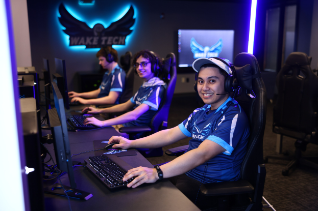 Gruppe von Menschen an einem Schreibtisch mit Computern, Kopfhörern und strahlenden Gesichtern; Schreibtisch mit Monitoren, Tastaturen und Mäusen; Hintergrund enthält ein Logo und eine Textwand, mit Stühlen rechts.