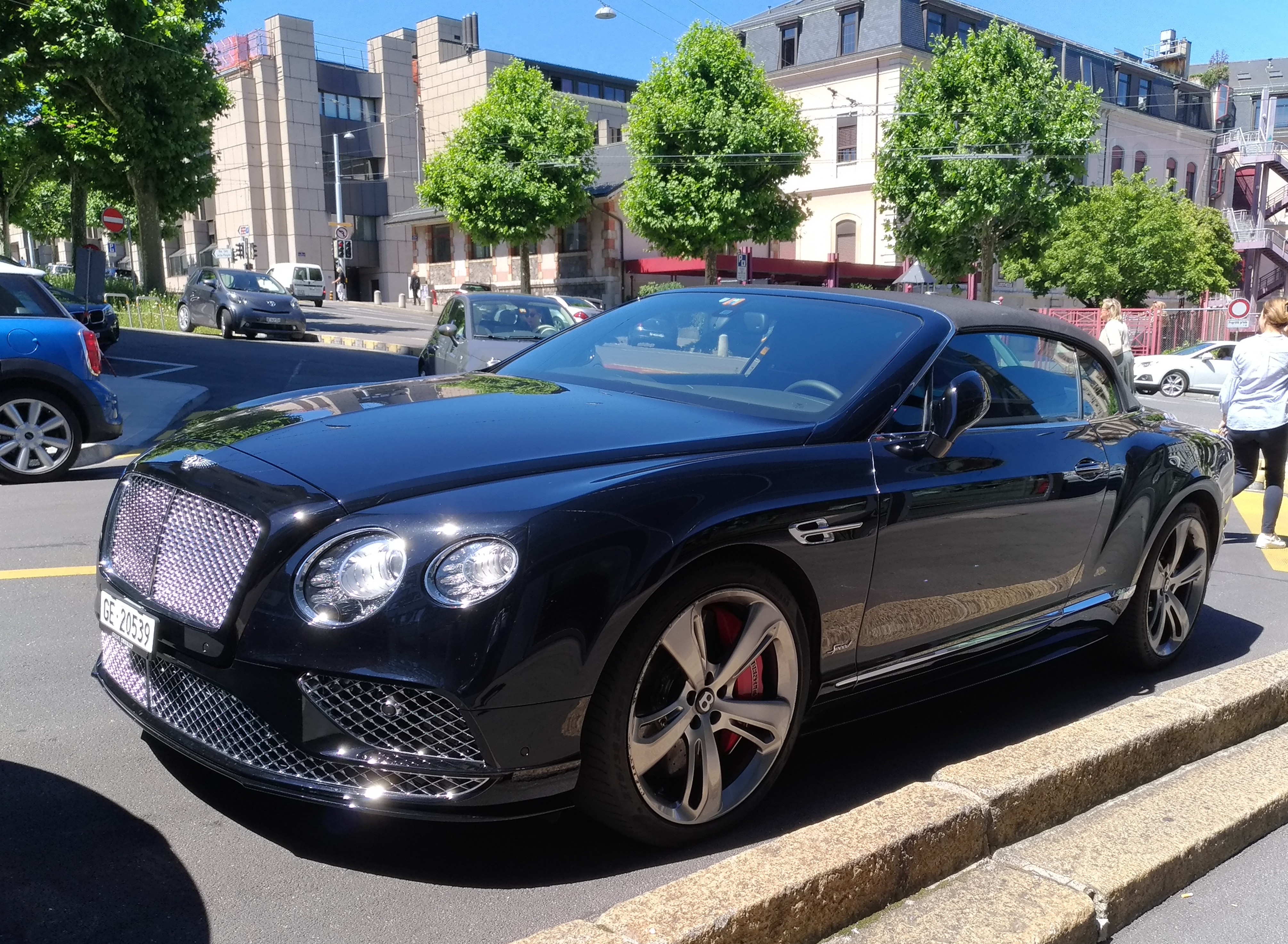 Ein Bentley Continental GTC V8 S fährt auf einer Straße, umgeben von Menschen, Bäumen, Gebäuden mit Fenstern, Laternenmasten und einem klaren blauen Himmel.