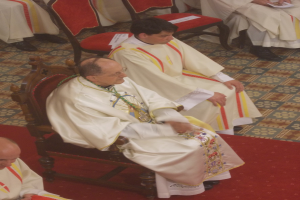 Eine Gruppe von Priestern in weißen Gewändern sitzt auf Stühlen auf einem roten Teppich in einer Kirche, einige halten Papiere.