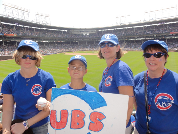 Drei Damen mit Kappen und Schutzbrillen, ein Junge mit Brille und Kappe, der einen Ball und ein Schild hält, stehen in einem Stadion mit Publikum und Feld dahinter, unter einem Himmel mit einigen Lichtern.